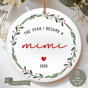 May include: A round white ceramic ornament featuring a green botanical wreath with red berries. The text reads THE YEAR I BECAME A mimi in black and red script, with a small red heart and the year 2026 printed below, suspended by a jute twine loop.