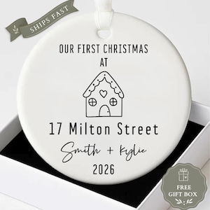 May include: A round white ceramic ornament featuring black text that reads OUR FIRST CHRISTMAS AT, 17 Milton Street, Smith + Kylie, and 2026. A black line drawing of a gingerbread house with a heart is centred above the address.