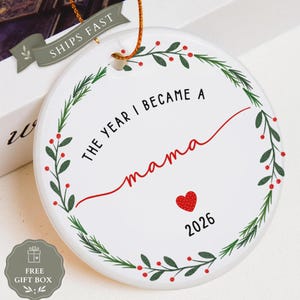 May include: A round white ceramic ornament featuring a green leaf and red berry wreath design. The text reads THE YEAR I BECAME A mama in black and red script, with a small red heart and the year 2026 printed below. It hangs from a gold string.