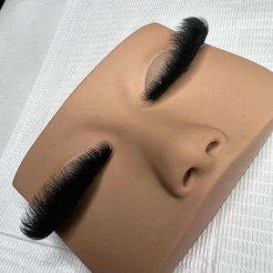 Op de afbeelding: Een beige mannequin-gezicht met lange, dichte, zwarte wimperextensions op beide gesloten oogleden. De wimpers zijn dik, hebben een uniforme lengte en creëren een dramatische, volle uitstraling langs de bovenste wimperrand van het model.
