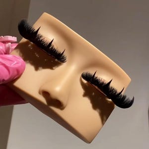 Pode incluir: Um manequim de treino facial em silicone bege com dois conjuntos de pestanas postiças pretas, longas e volumosas, aplicadas nas linhas das pestanas superiores. A placa é retangular com um nariz moldado e olhos fechados para treino.