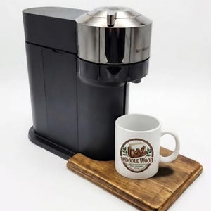 May include: A black coffee machine with a metallic silver top section sits next to a white ceramic mug featuring a brown logo that reads WOODLE WOOD. The items are placed on a rectangular wooden board with a natural grain and dark brown finish.