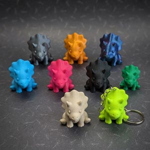 May include: A collection of nine small triceratops dinosaur figurines in various colors including gray, orange, blue, pink, black, green, beige, and lime green. One lime green triceratops is attached to a silver metal keychain ring.