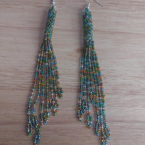 May include: A pair of long, dangling earrings featuring a dense, woven top section that transitions into multiple cascading strands of small, multi-colored glass beads in shades of emerald green, deep blue, bronze, and clear, finished with silver hooks.