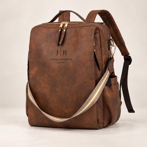 May include: A brown backpack featuring a top handle, adjustable shoulder straps, and gold-tone zips. The front is personalised with the initials JR and the name JENNA ROBERTS. A detachable, striped shoulder strap in white, grey, and gold is attached.
