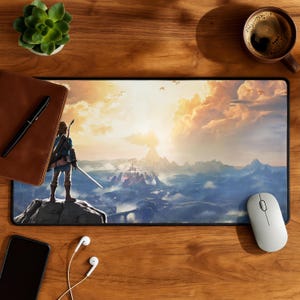 May include: A rectangular desk mat featuring a printed illustration of a character standing on a rocky cliff overlooking a vast landscape with mountains, a distant castle, and a bright sunset sky with clouds, all rendered in shades of blue, gold, and orange.