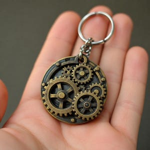 Può includere: Un portachiavi circolare in metallo color bronzo caratterizzato da molteplici ingranaggi interconnessi di varie dimensioni. L'oggetto include un anello in metallo argentato fissato tramite una piccola catena, dal design meccanico.