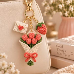 May include: A gold-toned metal keychain featuring three crocheted charms: a cream-colored envelope with a red heart, a red strawberry with green leaves, and a bouquet of pink tulips wrapped in cream yarn with a pink bow, all attached to a gold clasp.