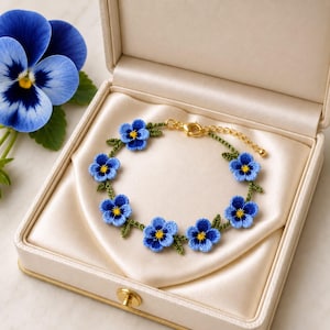 May include: A bracelet featuring seven small blue pansy flowers with yellow centers, connected by green thread stems and leaves. The piece includes a gold-toned metal lobster clasp and an adjustable chain link extension for secure fastening.
