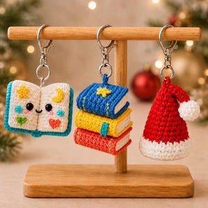 May include: Three crochet keychains hanging on a wooden stand. One is an open book with a smiling face, stars, and hearts. Another is a stack of three small books in blue, yellow, and red. The third is a red and white Santa Claus hat.