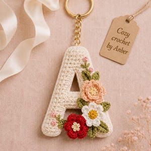 Puede incluir: Un llavero con la letra A tejida a ganchillo en color crema, con un anillo y cadena de metal dorado. La letra está decorada con pequeñas flores tejidas de colores en tonos melocotón, blanco y rojo, junto con hojas verdes y una etiqueta marrón con el texto Cozy crochet by Anber.