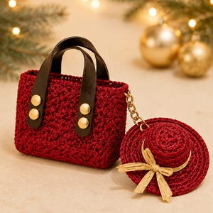 Könnte beinhalten: Eine dunkelrote gehäkelte Handtasche mit dunkelbraunen Ledergriffen, die mit goldfarbenen Metallnieten befestigt sind. An der Seite der Tasche ist eine passende, kleine, rote Häkelmütze mit einer hellbeigen Bastschleife an einer goldfarbenen Kette befestigt.
