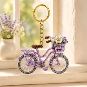 Puede incluir: Un llavero de bicicleta de ganchillo color lavanda con un asiento marrón, ruedas grises con radios blancos y una cesta delantera llena de pequeñas flores blancas y moradas. La bicicleta está unida a una anilla y cadena de metal dorado.
