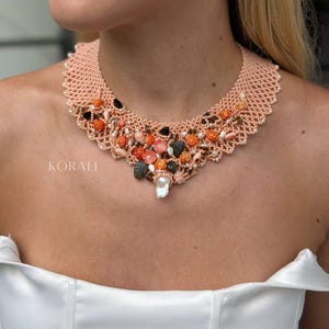 Puede incluir: Un collar de cuentas color melocotón con un tejido de cuentas de semillas, adornado con elementos florales tallados en tono coral, cuentas en forma de hoja verde oscuro y una perla barroca blanca central. La palabra KORALI aparece a la izquierda.