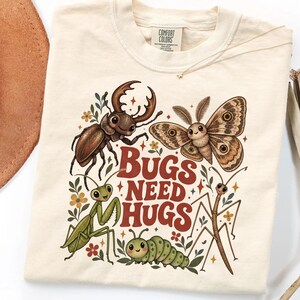 May include: A cream-colored t-shirt featuring a graphic print of a stag beetle, a moth, a praying mantis, a caterpillar, and a stick insect. The text "BUGS NEED HUGS" is printed in a bold, dark red font in the center, surrounded by small flowers.
