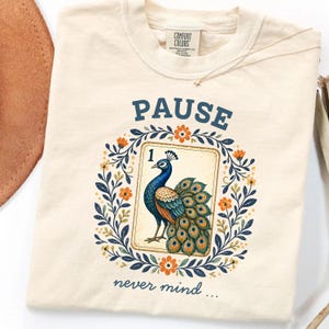 May include: A cream-colored t-shirt featuring a graphic of a peacock on a playing card labeled with the number 1. The peacock is surrounded by a floral wreath with blue leaves and orange flowers. The text reads PAUSE above and never mind below.