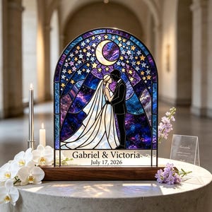 May include: An arched stained glass panel featuring a couple in wedding attire under a crescent moon and starry night sky. The base is dark wood with the text Gabriel & Victoria July 17, 2026. The glass displays deep blue and purple hues.