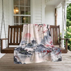May include: A soft throw blanket featuring a printed landscape illustration of a Japanese pagoda, misty mountains, a large pink sun, and blooming cherry blossom branches in shades of pink and red against a pale cream background.