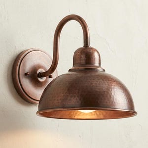 May include: A wall-mounted light fixture featuring a dome-shaped shade with a hammered copper finish. The lamp includes a curved arm extending from a circular backplate, with a warm light emitting from the underside of the metal dome shade.