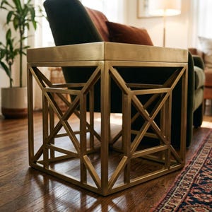 May include: A square side table with a metallic gold finish, featuring a geometric open-frame base constructed from intersecting diagonal and vertical bars. The table has a solid flat top surface and a sturdy, box-like structural design.