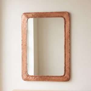 May include: A rectangular wall mirror featuring a wide, hammered copper frame with rounded corners. The reflective glass surface is centered within the metallic border, which displays a textured, uneven finish across its entire surface area.