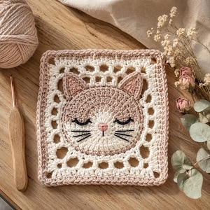 May include: A square crochet granny square featuring a central cat face design. The cat has a light beige and cream face with closed eyes, a pink nose, and black embroidered whiskers. The border is a cream and beige openwork crochet pattern.
