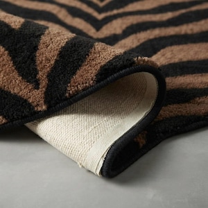 May include: A rectangular area rug featuring a black and brown zebra stripe pattern. The rug has a thick, textured pile surface with a black bound edge. One corner of the rug is folded over to reveal the light-colored, woven fabric backing underneath.