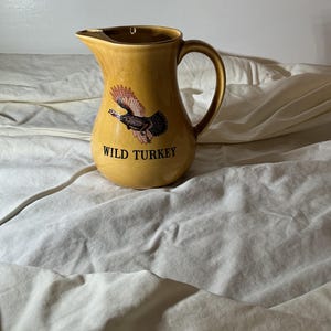 May include: A mustard yellow ceramic jug featuring a graphic illustration of a wild turkey in flight with spread wings. The text WILD TURKEY is printed in black capital letters below the bird. The jug has a curved handle and a pouring spout.