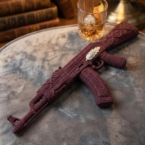 May include: A dark maroon crochet sculpture shaped like an assault rifle, featuring intricate textured stitching and a prominent silver-toned metallic leaf-shaped embellishment attached to the side of the barrel assembly.