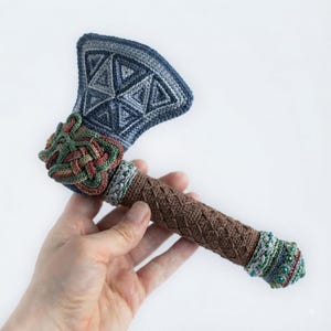 May include: A handcrafted crochet axe featuring a blue and grey triangular patterned blade, a brown textured handle with a cross-hatched design, and decorative green and red braided accents at the head and base with small green beads.