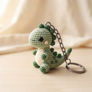 May include: A small, light green crocheted dinosaur keyring featuring a cream-coloured belly, dark green spikes along its back, and two black bead eyes. The figure is attached to a silver-toned metal chain and a circular split ring for keys.