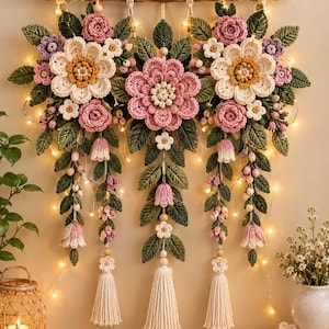 Op de afbeelding: Een wandhanger met gehaakte bloemen in roze, crème en paarse tinten met groene bladeren, opgehangen aan een houten tak. Het arrangement bevat drie hoofdgroepen van bloemen, hangende ranken en drie crèmekleurige kwastjes van garen.