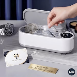 Könnte beinhalten: Ein weiß-grauer Ultraschallreiniger mit offenem Deckel, der Wasser und eine Metallbrille im Aviator-Stil enthält. Das Gerät verfügt über eine Digitalanzeige und einen Ein-Aus-Schalter. Eine Visitenkarte und ein goldenes Schild mit der Aufschrift Westfort Mart liegen daneben.