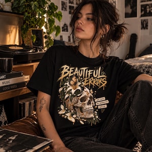Puede incluir: Una camiseta negra de manga corta con un estampado gráfico de un cráneo humano adornado con lirios blancos y una mariposa. El texto "BEAUTIFUL ERRORS" aparece en una fuente dorada, junto a recuadros que dicen "some chaos", "new perspective", "more me".