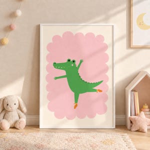 May include: A framed wall art print featuring a green crocodile character dancing on one leg. The crocodile is positioned against a large, scalloped-edge pink cloud shape on a cream background. The crocodile wears small orange slippers on its feet.