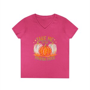 Pode incluir: Uma t-shirt fúcsia com decote em V apresentando um gráfico de três abóboras com bolinhas brancas e o texto "TAKE ME TO THE PUMPKIN PATCH" em letras laranja e azuis. A abóbora central é laranja, enquanto as duas abóboras laterais são brancas.
