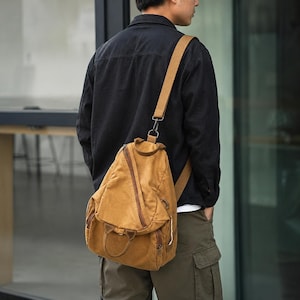 May include: A tan canvas sling backpack featuring a diagonal zippered front pocket, a top handle, and a single adjustable shoulder strap with a metal clip attachment. The bag has a structured, teardrop-shaped design with visible stitching details.