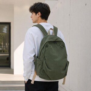 Può includere: Uno zaino in tela verde oliva scuro dotato di un ampio scomparto principale con chiusura a zip, una tasca frontale più piccola con cerniera e due tasche laterali. Lo zaino include spallacci beige regolabili e una maniglia superiore.