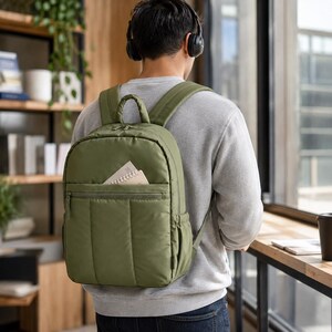 May include: An olive green backpack with a top handle, adjustable shoulder straps, and a front zippered pocket containing a small notebook. The backpack features a smooth, padded exterior with side pockets and matching green zipper pulls.