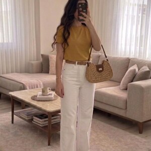 May include: A mustard yellow short-sleeved t-shirt paired with high-waisted white denim jeans, a thin brown leather belt, tan woven slide sandals, and a crescent-shaped straw handbag with a brown leather flap and a gold-tone metal clasp.