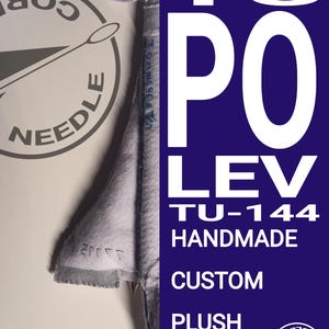 May include: A grey fabric plush toy shaped like a Tupolev TU-144 aircraft. The plush features stitched details and text on the side. A dark blue vertical panel on the right displays the white text TUPOLEV TU-144 HANDMADE CUSTOM PLUSH.