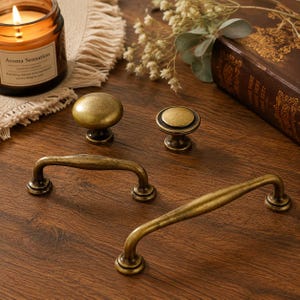 May include: A collection of four antique brass hardware pieces including two round knobs and two arched drawer pulls. The hardware features a weathered, textured finish with dark highlights, showcasing a classic design with circular bases.