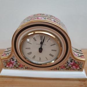 May include: A mantel clock with a cream-coloured face, black Roman numerals, and the text Wedgwood Quartz. The arched ceramic body features a floral pattern with pink and green tones, accented by gold trim on a white base with a gold-toned border.