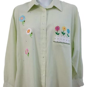 May include: A light green long-sleeve button-down shirt featuring embroidered floral appliqués and a chest pocket decorated with a white picket fence and three colourful flowers. The shirt has a collar and a vertical button placket down the centre.