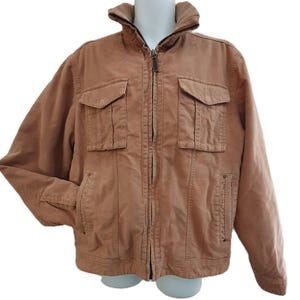 May include: A tan utility jacket featuring a full-length front zip, a high collar, and two chest pockets with button-down flaps. The jacket includes two vertical side pockets, long sleeves, and visible stitching details throughout the garment.
