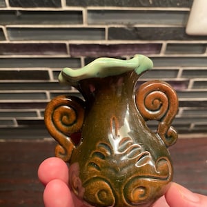 Può includere: Un piccolo vaso in ceramica con una smaltatura lucida marrone scuro e un bordo ondulato verde chiaro. Il vaso presenta due manici simmetrici a forma di voluta sui lati e un motivo a vortice in rilievo sulla parte anteriore con finitura liscia.