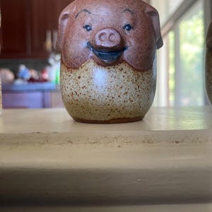 Può includere: Una statuetta di maiale in ceramica con un corpo arrotondato caratterizzato da una smaltatura bicolore. La parte superiore è di un marrone rossastro solido, mentre la sezione inferiore è color cuoio chiaro con macchie marrone scuro. Occhi, sopracciglia e bocca dipinti.