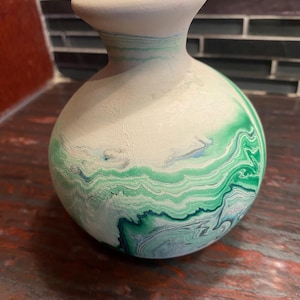 Könnte beinhalten: Eine kleine, runde Keramikvase mit engem Hals und ausgestelltem Rand. Die Oberfläche hat eine matte, cremeweiße Grundfarbe mit wirbelnden, marmorierten Mustern in verschiedenen Grüntönen, die von hellem Minzgrün bis zu dunklem Waldgrün reichen.