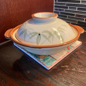 May include: A round ceramic donabe pot with a matching lid featuring a painted green bamboo leaf pattern. The pot has a cream-coloured base, a wide orange-brown rim, and two side handles. A small steam vent is visible on the lid's surface.