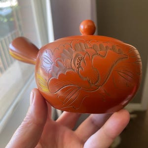 Peut inclure: Une théière orange brûlé avec une poignée latérale et un couvercle muni d'un bouton. Le corps de la théière est orné d'un motif floral sculpté complexe qui recouvre la surface avant, mettant en valeur des pétales et des feuilles en relief.